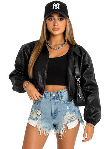 Woman wearing a black leather jacket, black crop top, ripped denim shorts, and a black cap with a white logo on a white background.